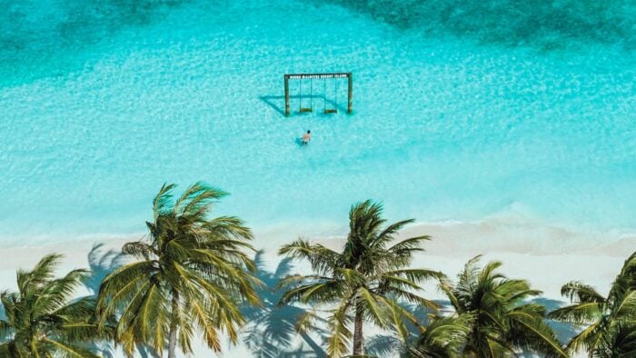 Aerial view of a guest walking toward the iconic swing at Meeru Maldives Resort Island, surrounded by turquoise water and palm trees on a white sandy beach.