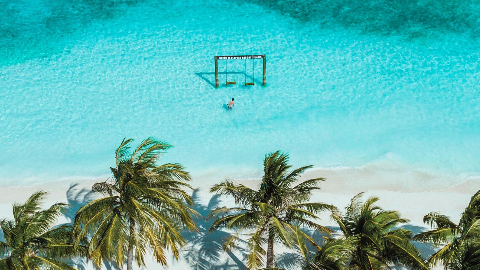 Aerial view of a guest walking toward the iconic swing at Meeru Maldives Resort Island, surrounded by turquoise water and palm trees on a white sandy beach.